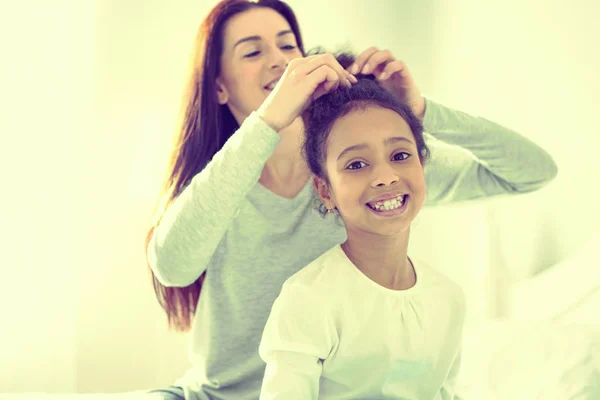Menina alegre recebendo seu cabelo feito por sua mãe carinhosa . — Fotografia de Stock