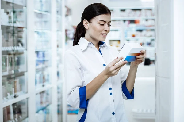 Χαριτωμένο μακρυμάλλης νεαρή γυναίκα σε ένα άσπρο παλτό που κρατά ένα κουτί με ένα νέο φάρμακο — Φωτογραφία Αρχείου