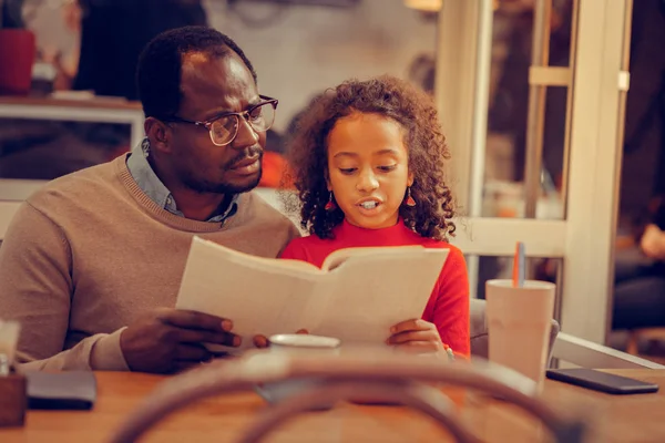 Curly smart daughter reading book on her own sitting near father — Stock Photo, Image