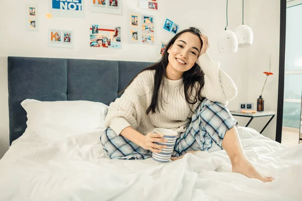 Alegre mujer de ojos oscuros bebiendo su té sentado en la cama — Foto de Stock