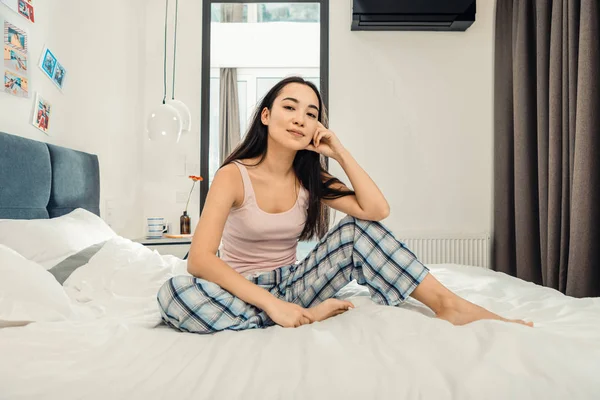 Mørkøyd kvinne med firkantede pysjbukser sittende på sengen – stockfoto