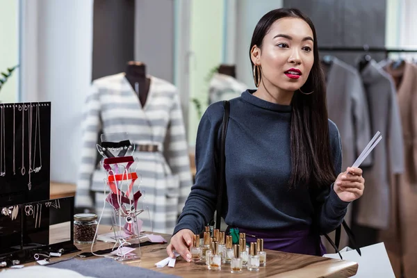 Dark-haired smiling asian lady in a dark-blue blouse testing fragrances