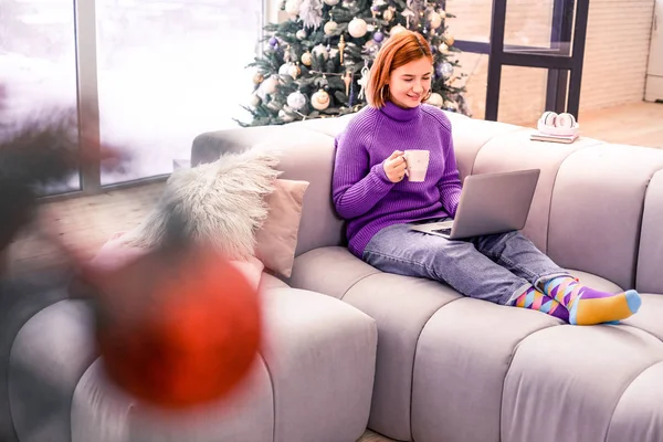 Pretty young red-haired beaming girl in a violet sweater reading emails — Stock Photo, Image