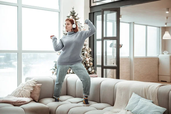 Egy szürke pulóver, tánc a kanapén sugárzó fiatal vörös hajú lány — Stock Fotó