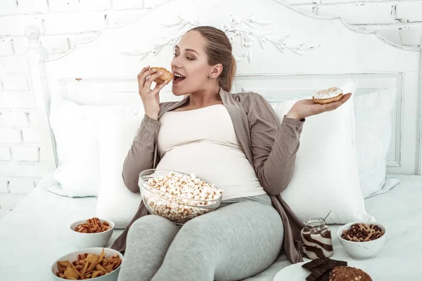 Cheerful thick woman lying in bed and filling herself with junk food
