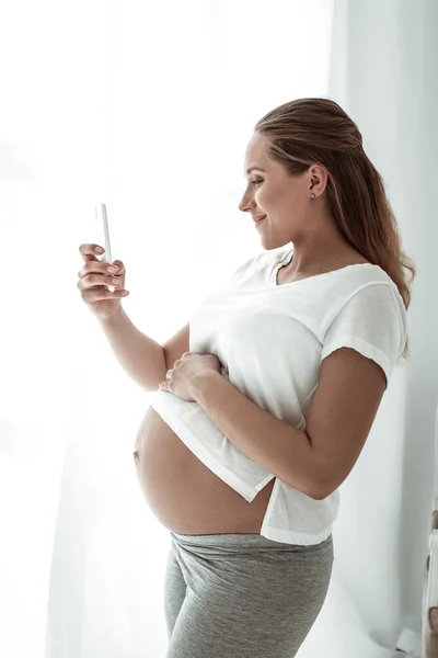 Сияющая красивая будущая женщина фотографирует свой беременный живот — стоковое фото