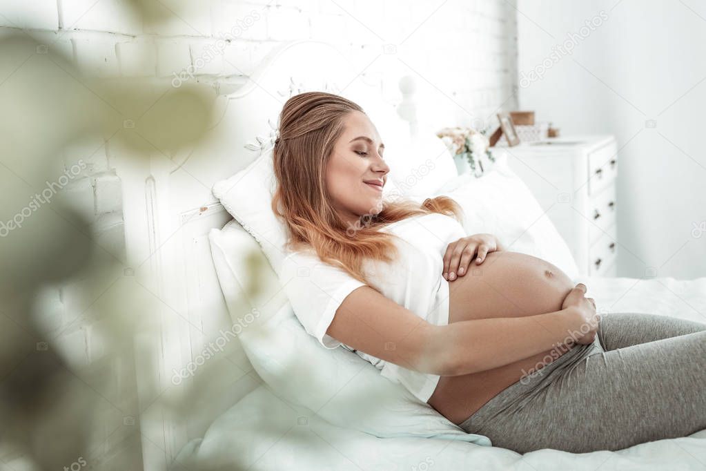 Tranquil long-haired pregnant woman touching her smooth stomach