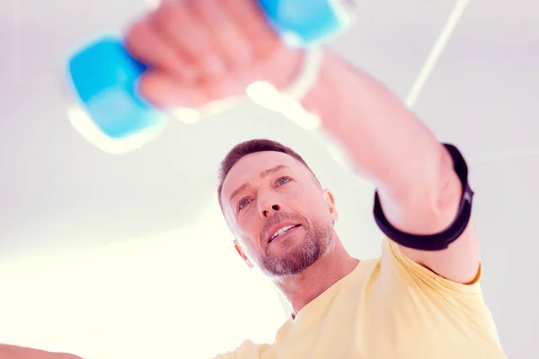 Bearded man wearing yellow shirt holding blue barbells while working on his body — Stock Photo, Image