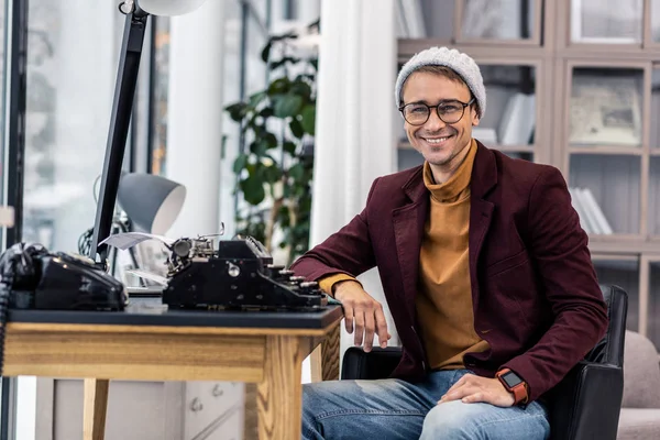 Well-groomed nifty smiling gentleman sitting at desk with typewriters