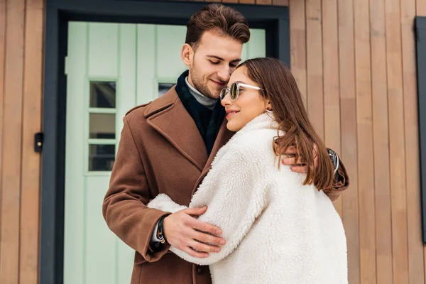 Loving wife wearing beige coat and glasses hugging her beloved — Stock Photo, Image