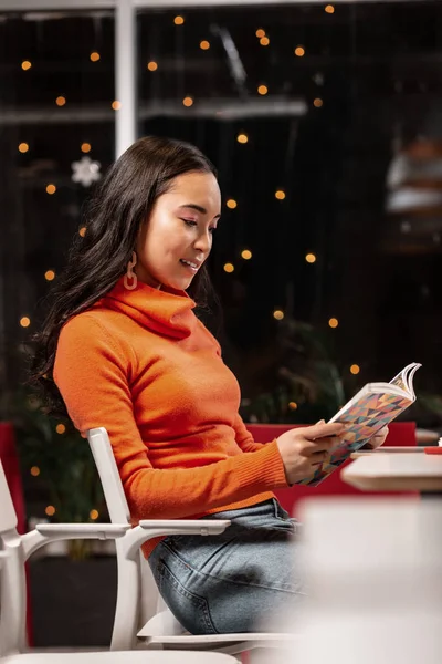 Asiatisches Mädchen liest Buch in gemütlichen atmosphärischen Café mit Lichterketten am Fenster. — Stockfoto