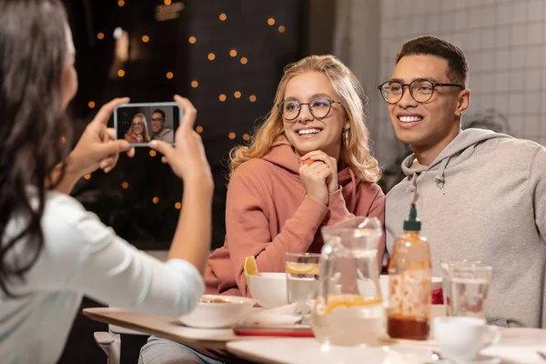 Девушка фотографирует знакомую пару в телефоне, пока они сидят и улыбаются . — стоковое фото