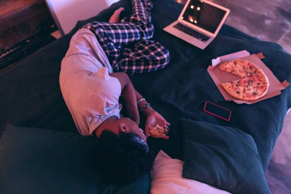 Уставший мальчик лежит на кровати с пиццей, телефоном, ноутбуком, пиццей и несколькими подушками . — стоковое фото