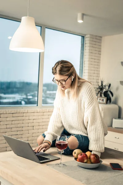 Uprzejmy Kobieta w jasne okulary siedzi na stole w kuchni i przy użyciu laptopa — Zdjęcie stockowe