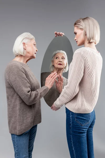 Nieuwsgierig oude dame aanraken oppervlak van het ovale spiegel in handen van haar dochter — Stockfoto