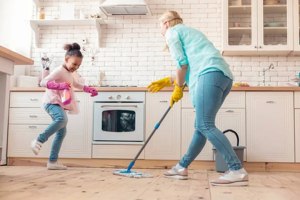 Lustiges Mädchen springt, während Mutter den Boden in ihrer Nähe wischt — Stockfoto