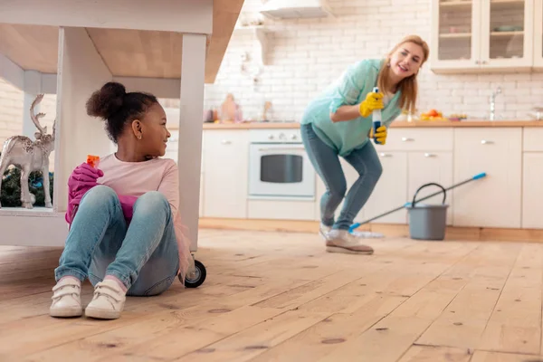 Cute curly daughter hiding under table while cleaning flat with mom — Stock Photo, Image