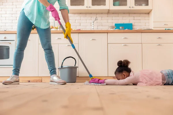 Funny curly girl lying on the floor while mother mopping the floor — Stock Photo, Image