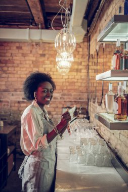 Young-adult beaming bewitching Afro-American bar employee holding towels at bar clipart