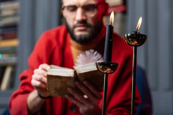 Bearded diviner wearing glasses reading old book sitting near candles — Stock Photo, Image