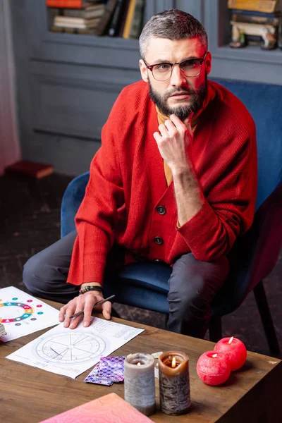 Темноволосий астролог у червоному кардигані сидить за столом зі свічками — стокове фото