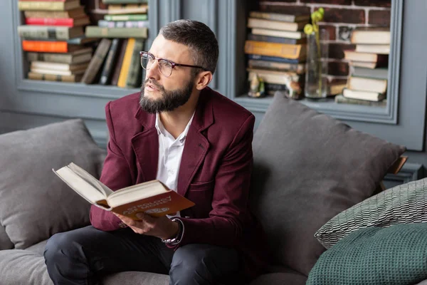 Handsome bearded man sitting on grey sofa and reading book — Stock Photo, Image