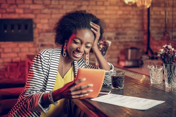 Bewitching Afro-American woman watching funny video on tablet at bar