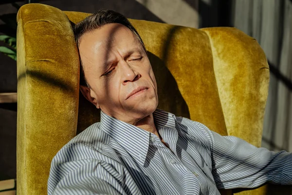 Short-haired upset man in striped light shirt lying on a yellow armchair — Stock Photo, Image