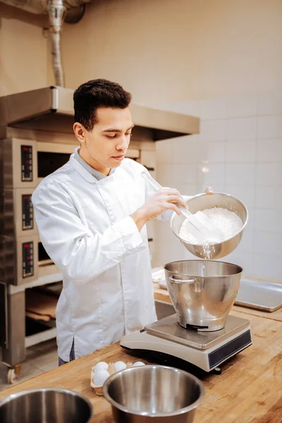 Dark-eyed professional baker adding some flour into bowl — Stock Photo, Image
