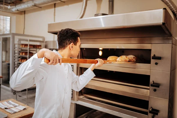 Молодой пекарь берет готовый хлеб из большой духовки на кухне — стоковое фото