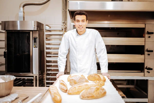 Jovem padeiro habilidoso de pé na cozinha perto da bandeja com pão — Fotografia de Stock