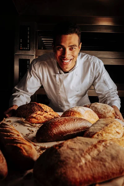 Baker sorrindo amplamente de pé perto da mesa com pão — Fotografia de Stock