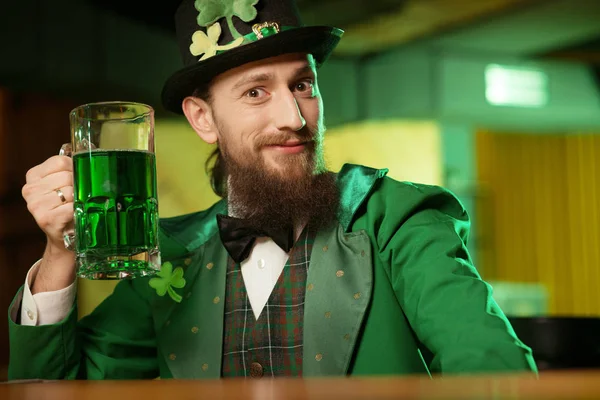 Σκούρα μαλλιά γενειοφόρος νεαρός άνδρας σε ένα leprechaun καπέλο με τριφύλλι, αυξάνοντας το ποτήρι του με πράσινο μπύρα — Φωτογραφία Αρχείου