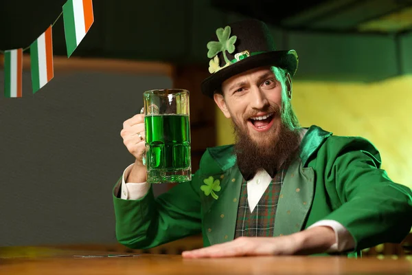 Jeune homme barbu aux cheveux bruns dans un chapeau de lutin avec du trèfle buvant de la bière verte — Photo