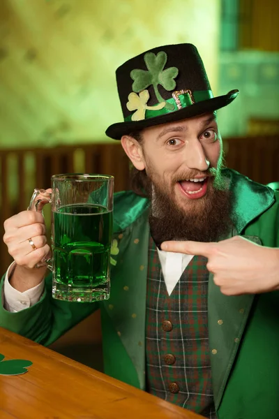 Giovane barbuto dai capelli scuri in un cappello leprecauno con trifoglio che punta al bicchiere con la birra — Foto Stock