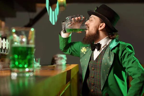 Dark-haired bearded young man in a green vest with shamrock drinking green beer — Stock Photo, Image