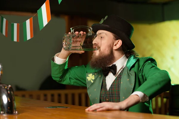 Темноволосый бородатый молодой человек в костюме лепрекона пьет зеленое пиво из большого стакана — стоковое фото