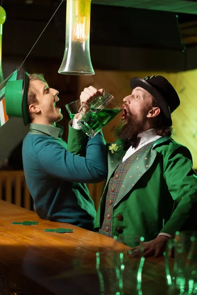 Jeune homme barbu aux cheveux bruns dans un chapeau de lutin et son ami aux cheveux blonds buvant de la bière verte — Photo