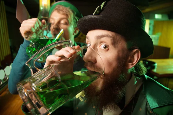 Fair-haired young man in a green hat and his bearded friend drinking lots of green beer — Stock Photo, Image