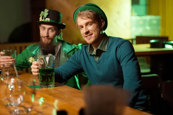Bel giovanotto dai capelli biondi con un cappello verde e il suo amico in costume da folletto seduto al bancone del bar — Foto Stock