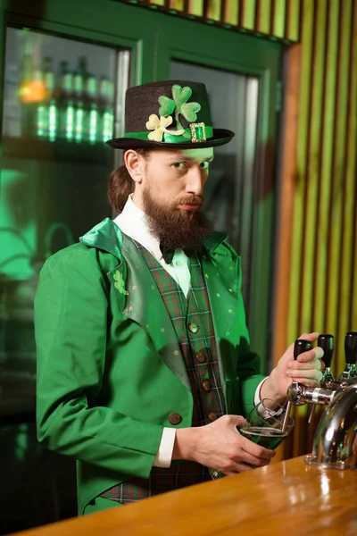 Jeune homme barbu aux cheveux foncés dans un chapeau de lutin versant de la bière verte dans le verre — Photo