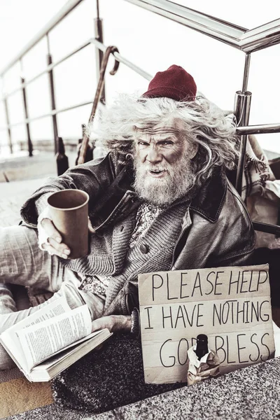 Pensive old almsman sitting and reading book while trying to earn some alms. — Stock Photo, Image