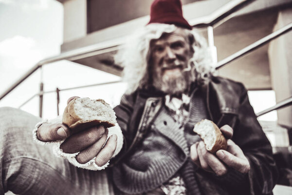 Portrait of old vagrant sitting and sharing his bun with stranger.