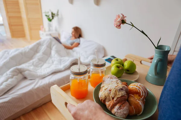 Esposa acostada en la cama mientras el marido trae bandeja con croissants y frutas — Foto de Stock