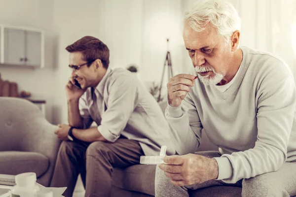 Old man taking pills while son indifferently having a phone call. — Stock Photo, Image