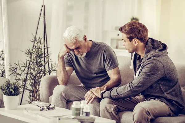 Young-adult short-haired son comforting elderly grey-haired upset father at living-room. — Stock Photo, Image