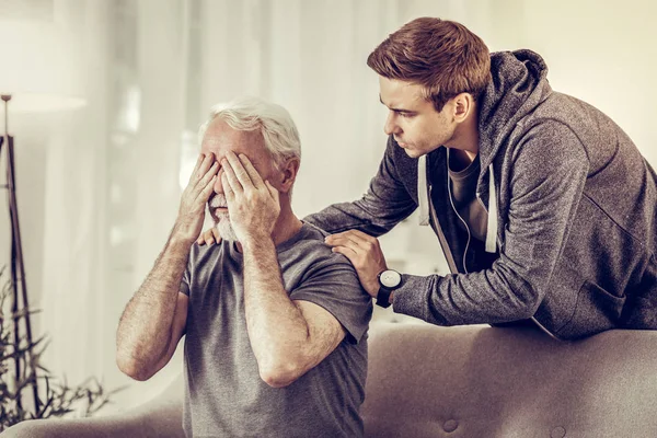 Молодой привлекательный расстроенный мужчина спокойно обнимает больного седого деда — стоковое фото