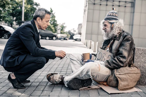 Pleasant short-haired rich man in costume supporting miserable homeless