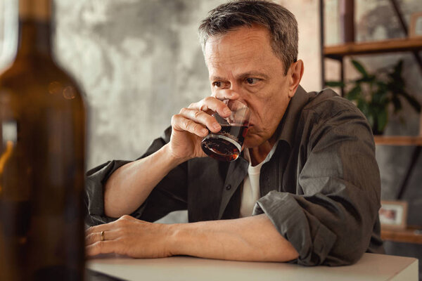 Apathetic grey-haired man sipping alcohol from glass