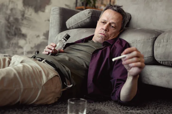 Apatyczny siwy mężczyzna leżąc na dywanie na sam na sam z butelki alkoholu — Zdjęcie stockowe
